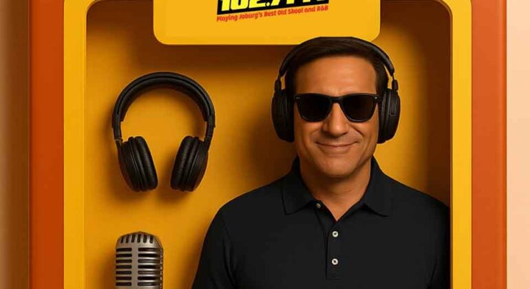 A man wearing headphones and sunglasses stands smiling inside a yellow radio booth with a microphone, headphones, and a HOT 102.7 FM sign above him.