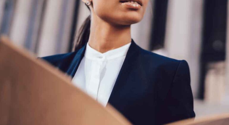 A confident woman in glasses and a business suit holds a large open folder, looking up thoughtfully outside a building with columns.