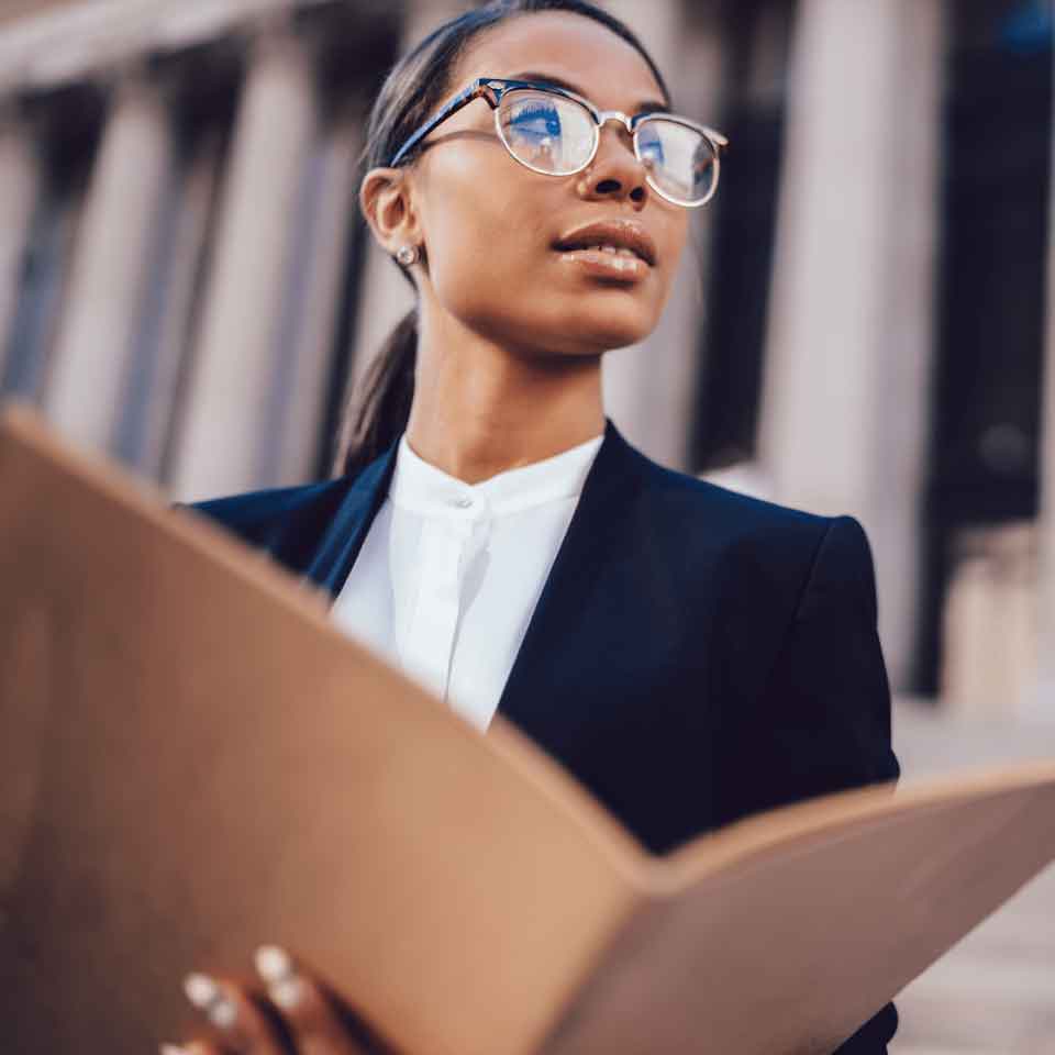A confident woman in glasses and a business suit holds a large open folder, looking up thoughtfully outside a building with columns.