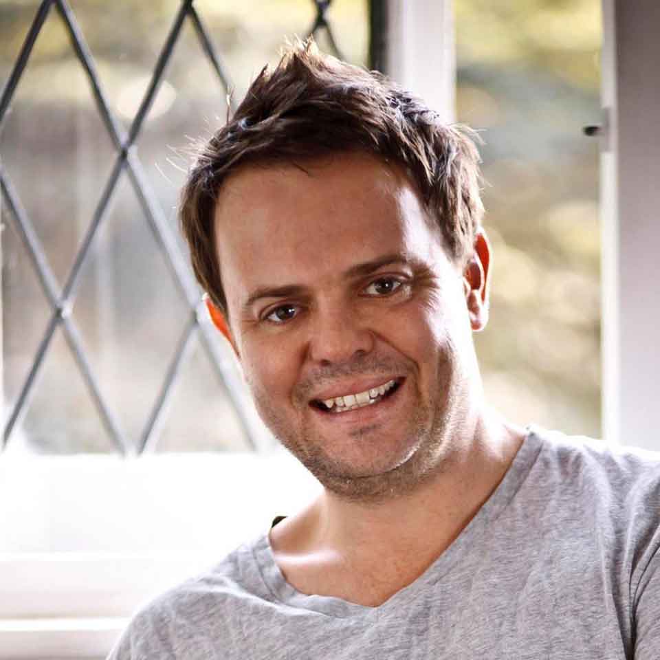 A man with short brown hair and a light beard smiles at the camera. He is wearing a light gray t-shirt and is sitting indoors near a window with a diamond-patterned frame.