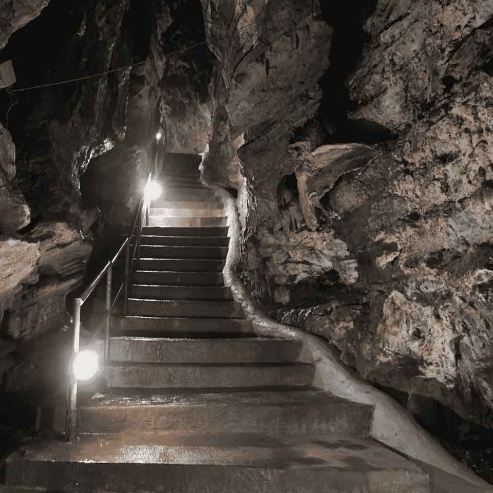 Stone steps with a metal handrail ascend through a dimly lit, rocky cave interior; lights along the rail illuminate the damp, uneven rock walls surrounding the staircase.