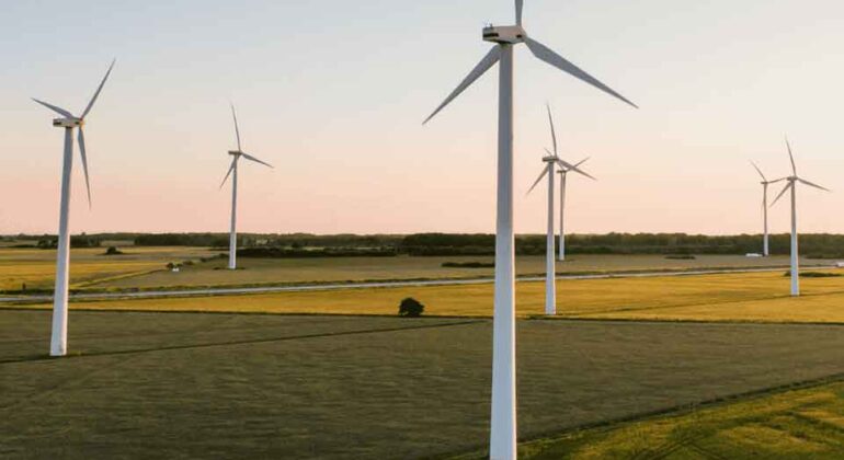 Wind turbines stand in a green field at sunset, generating renewable energy. The sky is clear with a warm hue, and the landscape is flat with scattered patches of trees and distant horizon.