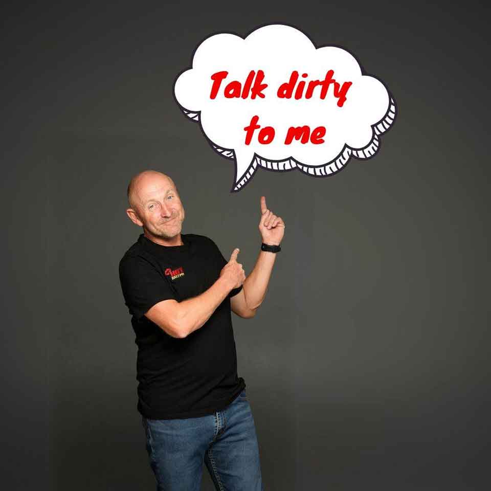 A bald man in a black t-shirt and jeans stands against a dark background, smiling and pointing up at a cartoon speech bubble that says, Talk dirty to me in red letters.