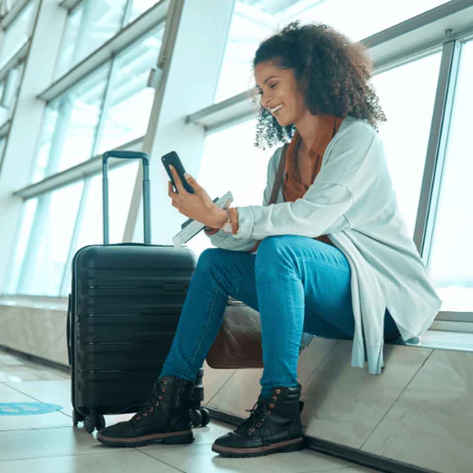 A woman sits in an airport terminal next to a suitcase, smiling while looking at her phone. She is casually dressed in jeans, boots, and a cardigan, with large windows and natural light in the background.