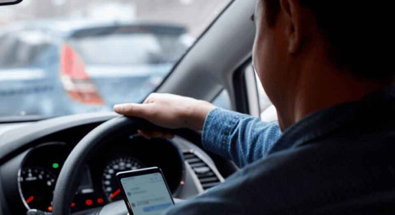 A person sits in the driver’s seat of a car, holding the steering wheel with one hand and looking at a smartphone in the other hand, while the car is stopped in traffic.