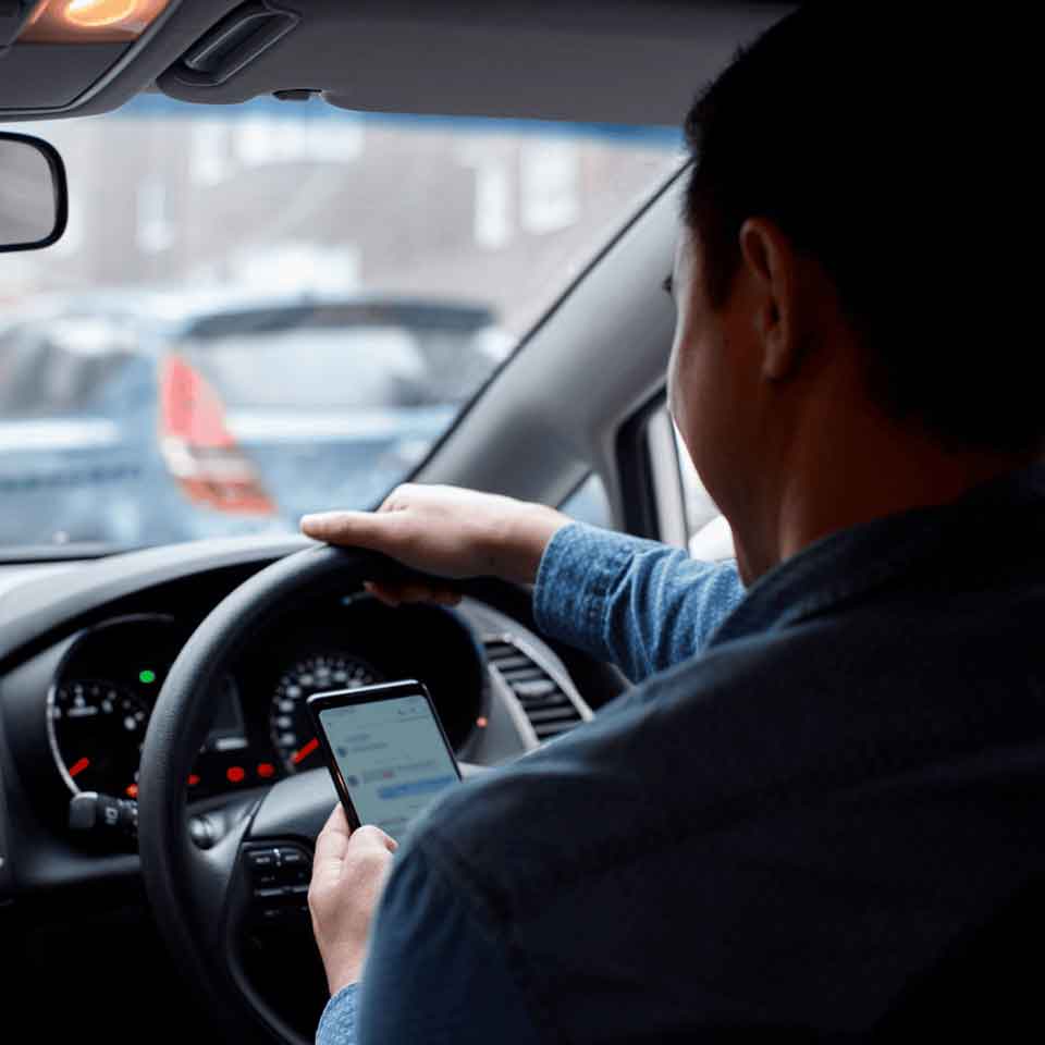 A person sits in the driver’s seat of a car, holding the steering wheel with one hand and looking at a smartphone in the other hand, while the car is stopped in traffic.