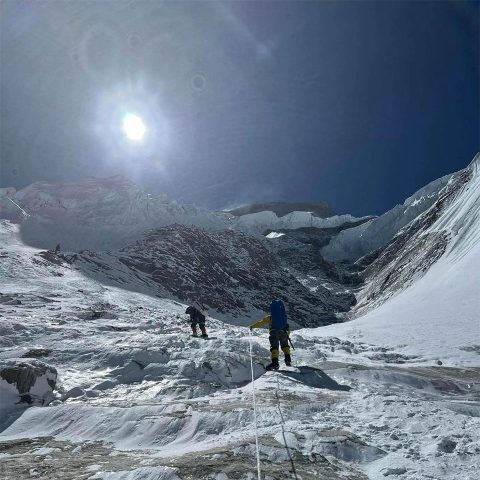 Two climbers, dressed in winter gear, ascend a snowy, sunlit slope on Annapurna Mountain under a bright blue sky, surrounded by steep, icy ridges and rugged terrain.