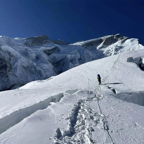 A lone climber in winter gear ascends a snow-covered, steep slope on Annapurna Mountain, following a trail of footprints. Jagged ice and snow formations tower in the background under a clear blue sky.