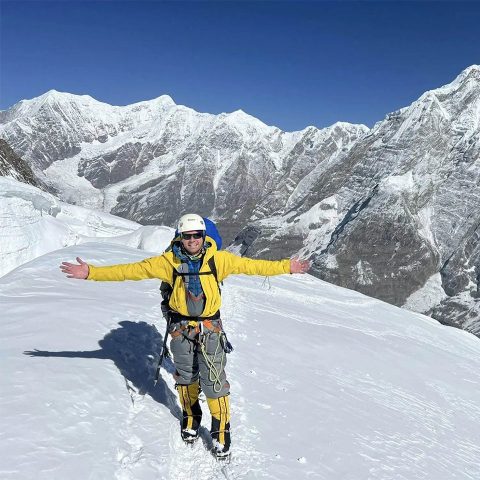 A mountaineer in a yellow jacket and climbing gear stands on a snowy slope with arms outstretched, surrounded by the towering, snow-covered peaks of Annapurna Mountain under a clear blue sky.