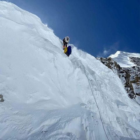 Two climbers in winter gear scale a steep, snowy Annapurna Mountain slope using ropes, with a bright blue sky and a snow-covered peak visible in the background.