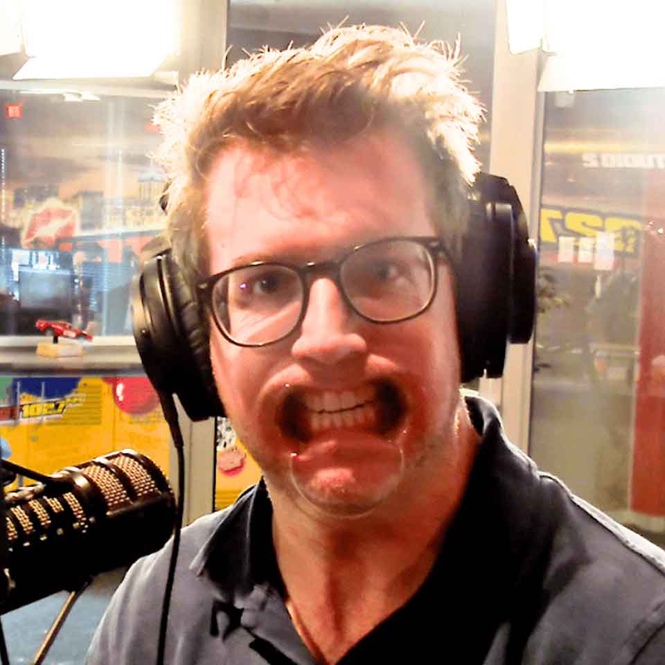 A man with glasses and headphones sits in front of a microphone, pressing his face against a clear surface, distorting his mouth and cheeks in a humorous way.