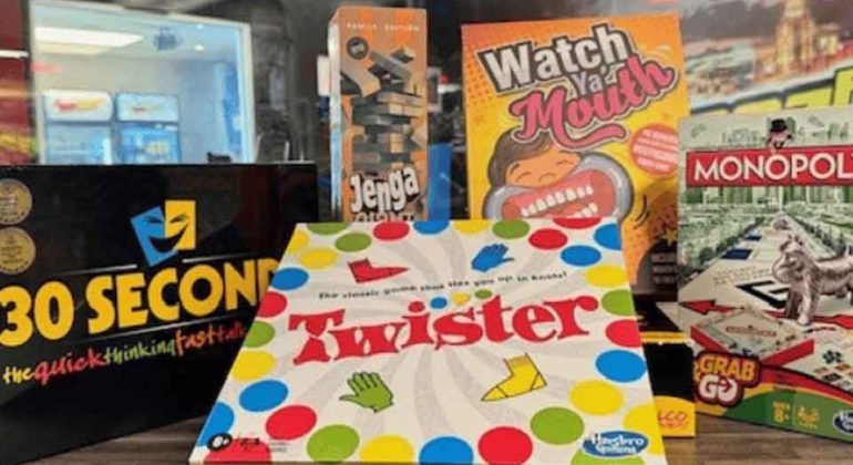 A display of board games on a wooden surface, including 30 Seconds, Jenga, Watch Ya Mouth, Monopoly, Grab & Go, and Twister, with shelves and store merchandise visible in the background.