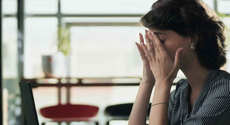 A woman sitting at a desk with a laptop, covering her face with her hands in frustration or exhaustion. A red mug is on the table, and the background shows large windows with natural light.