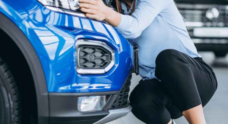 A smiling woman crouches next to a blue car, hugging its front and resting her head near the headlight inside a showroom.