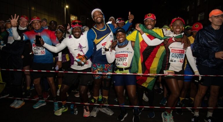 A group of enthusiastic marathon runners, some holding a Zimbabwean flag, smile and pose for the camera at the starting line during a nighttime Comrades Marathon race.
