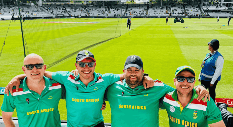 Four smiling men wearing South Africa Proteas cricket shirts pose arm-in-arm at a cricket stadium, with bright green grass, equipment, and other people visible in the background.