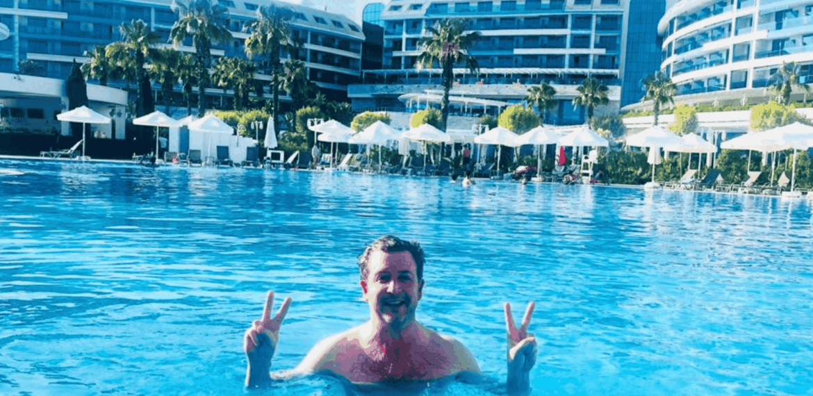 Simon Hill stands in a large outdoor swimming pool in Turkiye, smiling and holding up double peace signs with both hands. In the background are modern resort buildings, palm trees, and white poolside umbrellas.