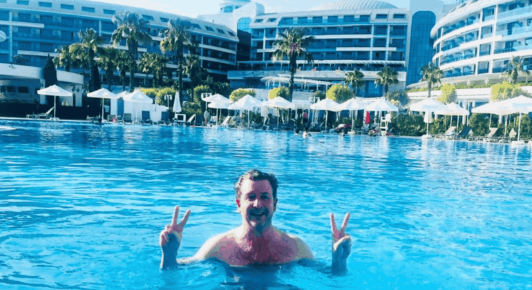 Simon Hill stands in a large outdoor swimming pool in Turkiye, smiling and holding up double peace signs with both hands. In the background are modern resort buildings, palm trees, and white poolside umbrellas.