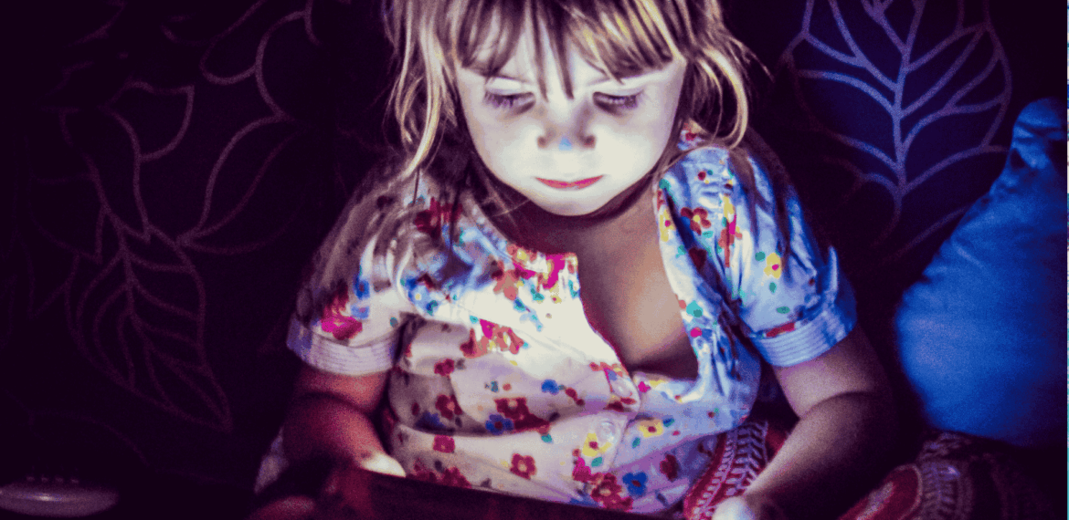A young child in colorful pajamas sits in dim lighting, illuminated by the glow of a tablet screen, with a patterned cushion and dark leaf-patterned background behind them.