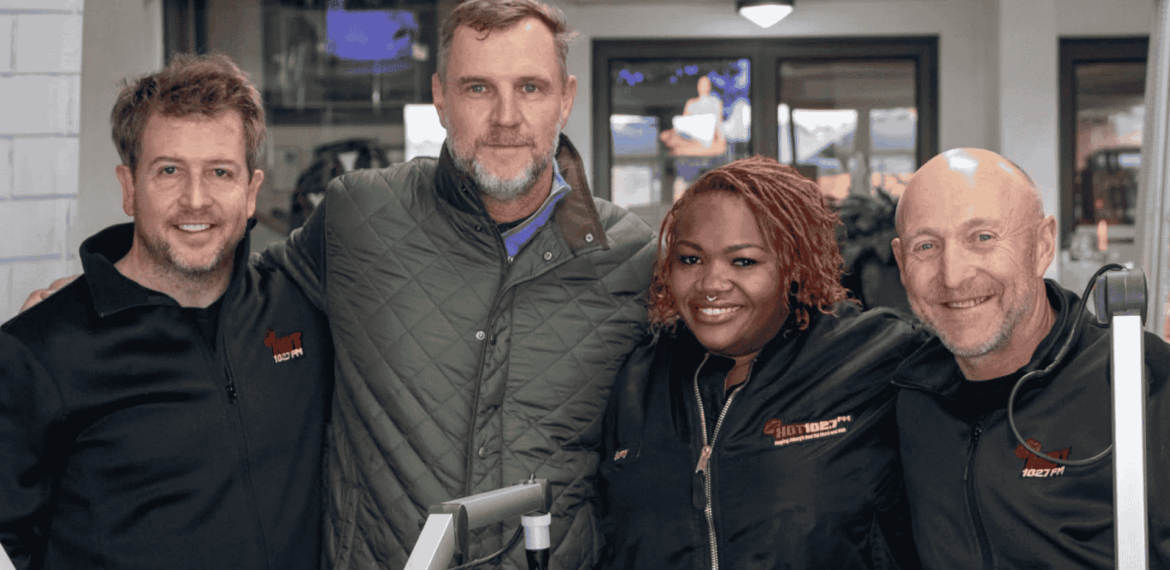 Four people stand closely together, smiling at the camera. Two wear black jackets with Sunshine Tour logos, one sports a green quilted jacket, and another wears a black shirt. Equipment and a TV screen are visible in the background.