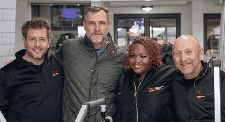 Four people stand closely together, smiling at the camera. Two wear black jackets with Sunshine Tour logos, one sports a green quilted jacket, and another wears a black shirt. Equipment and a TV screen are visible in the background.