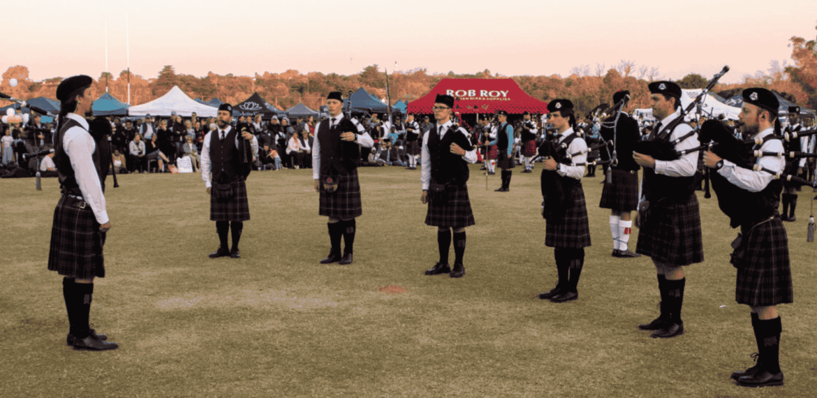 A group of people in kilts and uniforms play bagpipes in a circle on a grassy field during a Highland gathering, with tents and a crowd of onlookers in the background under a clear sky.