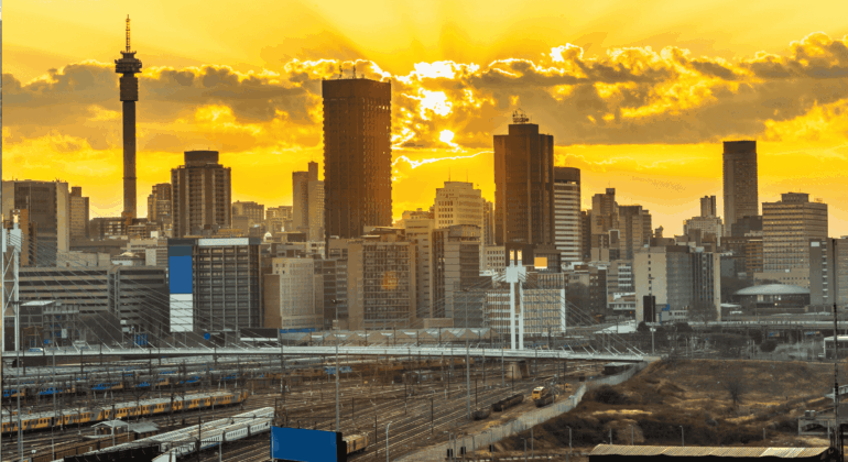 City skyline with tall buildings silhouetted against a dramatic yellow and orange sunset, with train tracks and railway infrastructure in the foreground—a striking view symbolizing efforts to rebuild Johannesburg.