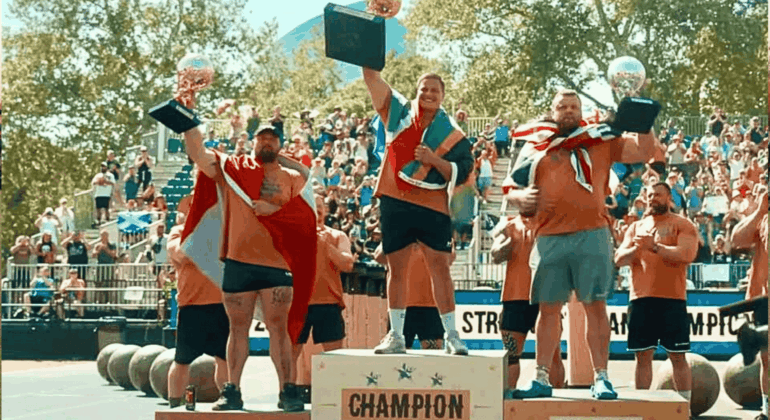 Three men, including Ryno Nel, stand on a winners’ podium at a strongman competition, holding trophies and draped in flags. The central champion raises his trophy high as a cheering crowd and large weights fill the background.