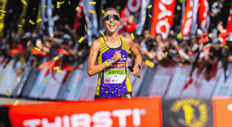 A female runner, Gerda Steyn, smiles as she crosses the finish line, surrounded by cheering crowds and confetti. She wears sunglasses, a purple and yellow athletic outfit, and a race bib reading Gerda.