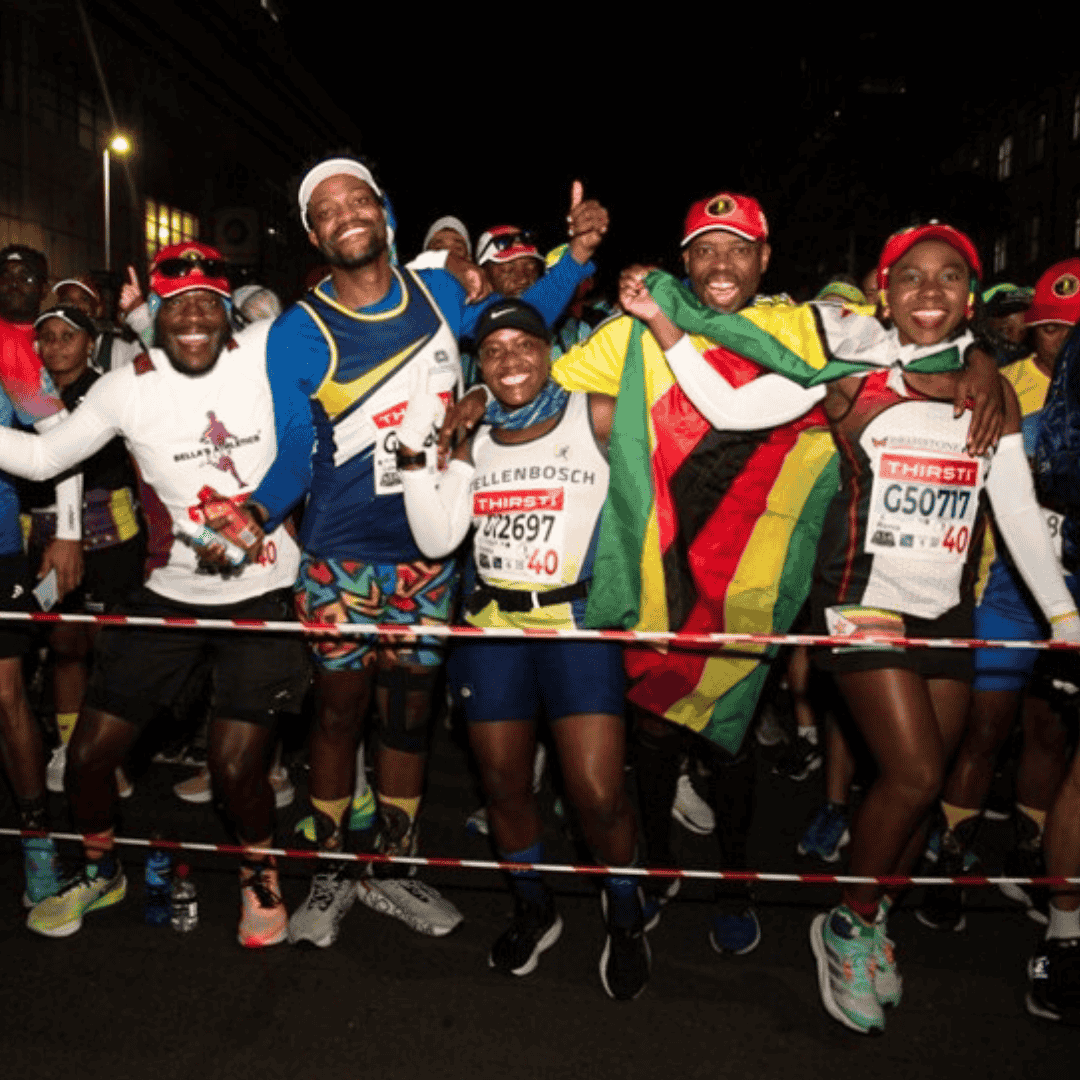 A group of smiling marathon runners pose together at the starting line of the Comrades Marathon, holding a Zimbabwean flag and wearing colorful athletic gear and race bibs, with more runners visible in the background.