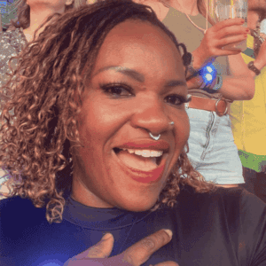 A person with curly hair and a nose ring smiles widely at the camera in their dark top. In the background, others—perhaps fellow Beyhive fans—stand holding drinks as sunlight illuminates this lively outdoor social event.