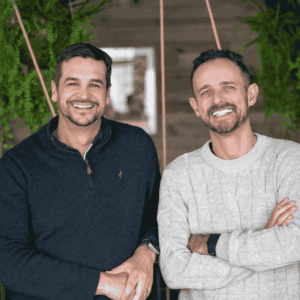 Two men standing side by side, both smiling at the camera. Indoors with green plants hanging in the background, they look like friends ready to expat explore new adventures together—one in a dark sweater, the other in light with arms crossed.
