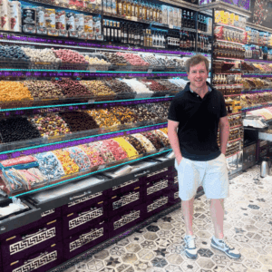 A man in a black shirt and white shorts stands smiling inside a colorful market in Turkiye, surrounded by shelves filled with various spices, dried fruits, and bottles of oil. The floor features a vibrant geometric tile pattern.