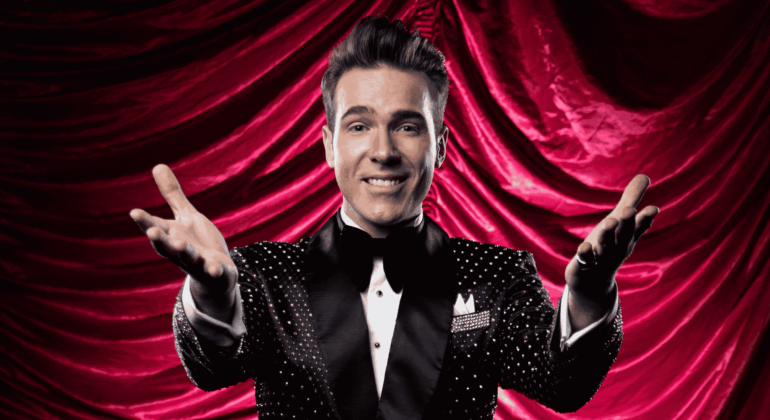 A smiling man in a sparkly black tuxedo with a bow tie stands in front of dramatic red curtains, extending both arms forward in a welcoming gesture, capturing the excitement of a Broadway performance.