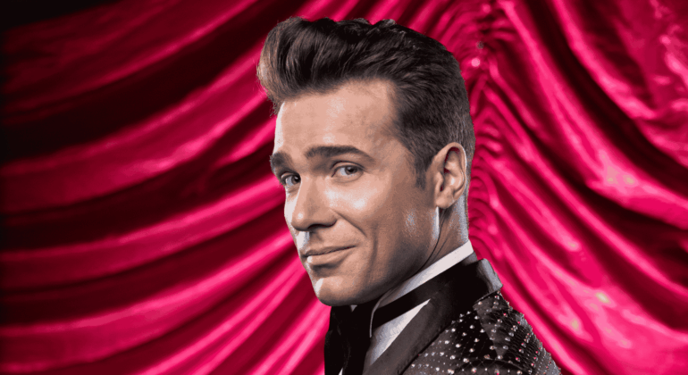 Jonathan Roxmouth, in a sparkling black tuxedo, smiles confidently over his shoulder against a dramatic, red velvet curtain backdrop.