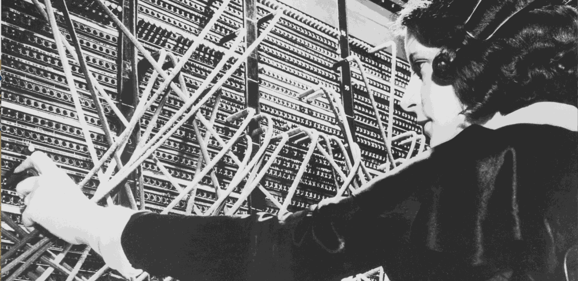 A woman operates a vintage telephone switchboard, connecting calls by inserting cables into various ports. Amid a tangle of wires, her focused demeanor recalls the playful coordination found in a bustling schoolyard.