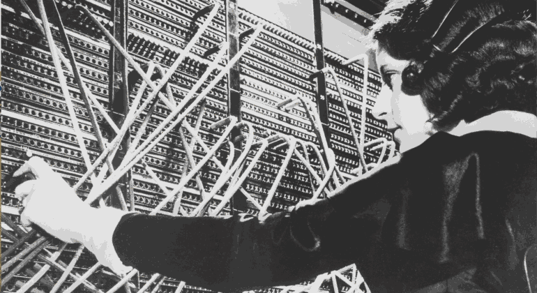 A woman operates a vintage telephone switchboard, connecting calls by inserting cables into various ports. Amid a tangle of wires, her focused demeanor recalls the playful coordination found in a bustling schoolyard.