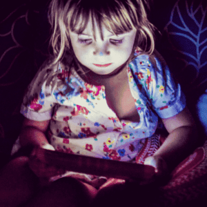 A young child with light hair sits in a dimly lit room, illuminated by the glow of a tablet screen, and wears a colorful floral-patterned shirt while looking intently at the device.