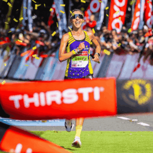 A smiling female runner, reminiscent of Gerda Steyn, wearing sunglasses and a purple-and-yellow outfit crosses the finish line as confetti falls and a cheering crowd celebrates her victory.