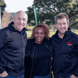 Three people stand close together outdoors, smiling at the camera on their Sunshine tour. They all wear black jackets, with green trees and grass in the background under a clear sky.