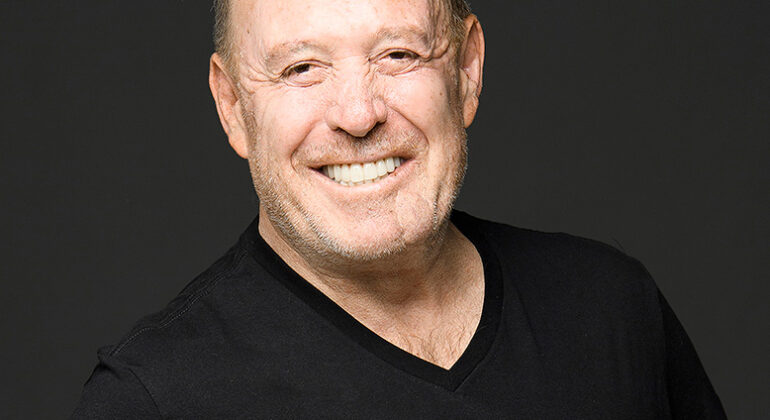A smiling man in a black t-shirt with the HOT 102.7 FM logo stands against a plain dark background, ready to broadcast live from a special memorial service.