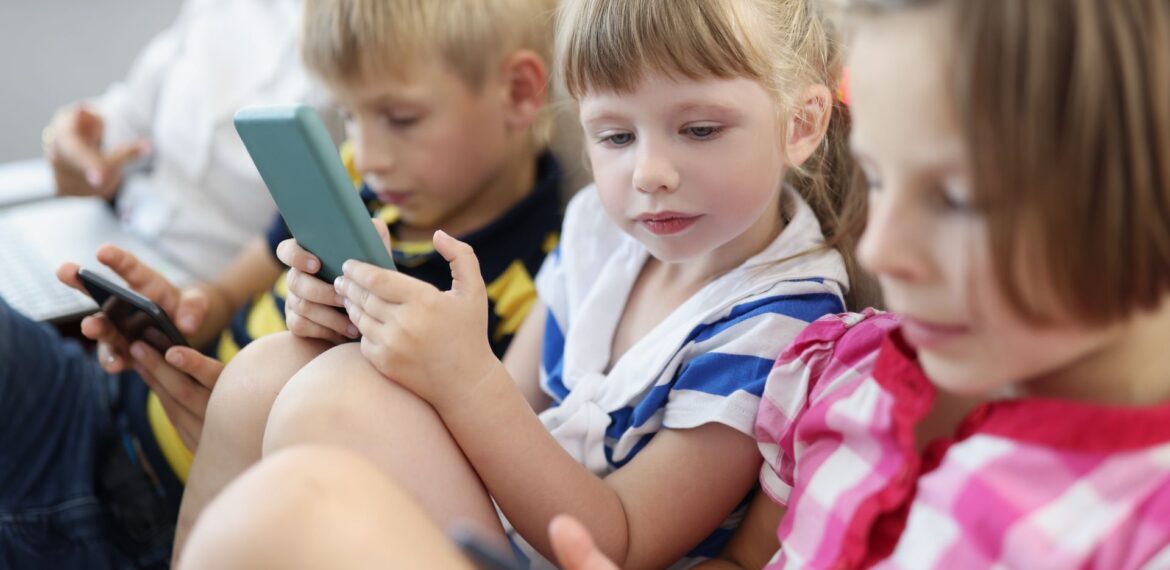 Three young children sit closely together in a bright indoor setting, each focused on a smartphone or tablet screen. Dressed in casual striped clothing, they appear fully engaged with their devices, absorbed in their screen time.