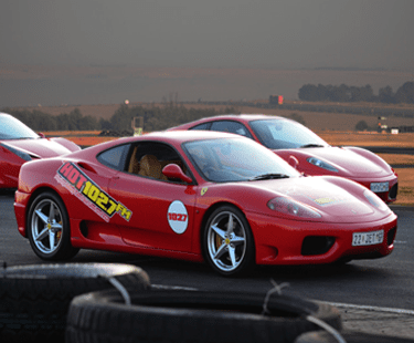 A red sports car with HOT 102.7 FM decals is parked on a racetrack, with another red sports car and a scenic landscape visible in the background. Tires are stacked in the foreground.