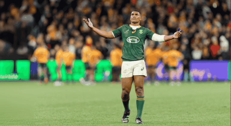 A rugby player in a green jersey stands with arms outstretched on a field, looking upward. The background shows blurred players in orange jerseys and a crowd in the stands.