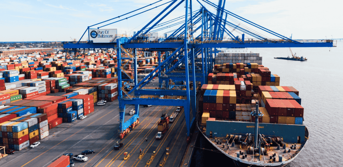 A large cargo ship loaded with colorful shipping containers is docked at the Port of Baltimore, with cranes and stacks of containers visible along the waterfront.