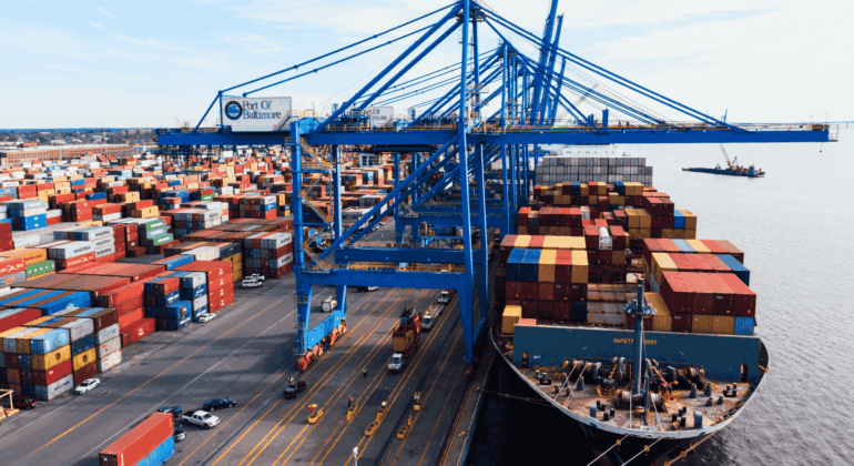 A large cargo ship loaded with colorful shipping containers is docked at the Port of Baltimore, with cranes and stacks of containers visible along the waterfront.