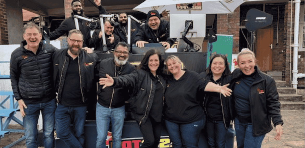A group of people wearing black jackets stand and smile in front of an outdoor radio station setup, with some people sitting or standing behind a DJ booth under a white umbrella. Everyone looks happy and cheerful.