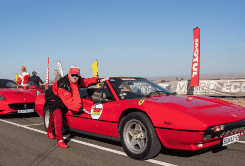A person in red racing gear poses next to a vintage red Ferrari on a racetrack, with another red Ferrari and people in similar attire in the background. Sponsor banners and clear skies complete the scene.