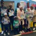 A group of children and teens stand outdoors, holding certificates and smiling for a photo. They are dressed warmly in jackets and hats, with colorful walls behind them. An adult’s hand is visible, taking the picture.