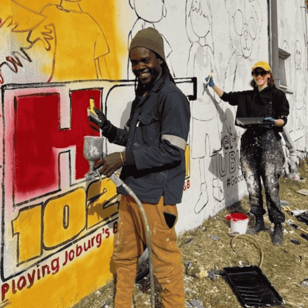 Two artists paint a mural on an outdoor wall. The man in front smiles while painting near a large red and yellow HOT 102.7 logo. The woman behind him paints outlines of cartoon figures. Painting supplies are on the ground nearby.